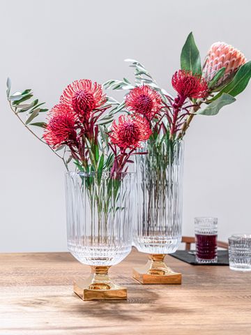 Two NACHTMANN Minerva vases with golden foot, filled with a bouquet of large red flowers on a light wooden table. In the background are a longdrink glass and a tumbler of the NACHTMANN series Ethno.<br/>