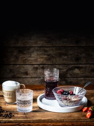 The NACHTMANN Ethno series on a wooden table. The hot beverage mug is filled with cappuccino, the tumbler with mineral water. The longdrink glass, filled with berry juice and the bowl, filled with a crunchy berry muesly, are standing on a white serving tray.<br/>