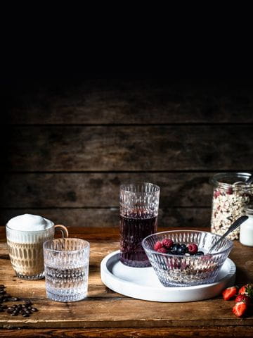 The NACHTMANN Ethno series on a wooden table. The hot beverage mug is filled with cappuccino, the tumbler with mineral water. The longdrink glass, filled with berry juice and the bowl, filled with a crunchy muesly, are standing on a white serving tray. In the background a jar with cereals and milk.<br/>