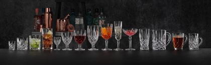 La collezione NACHTMANN Noblesse allineata su un bancone da bar nero, sullo sfondo bottiglie e attrezzature da bar. Il tumbler Noblesse, il bicchiere da longdrink, il bicchiere da cocktail e la coupette sono riempiti con diversi cocktail. Il calice da vino è pieno di vino rosso, la tazza per bevande calde è piena di tè. Gli altri bicchieri sono vuoti.<br/>