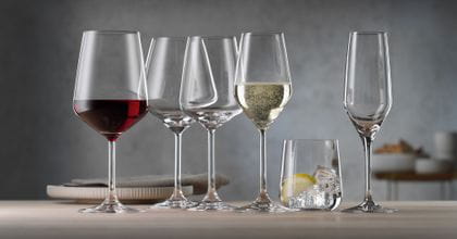 Das gefüllte SPIEGELAU Style Rotweinglas neben dem leeren Burgunderglas, dem Weißweinglas, dem gefüllten Champagnerglas, dem mit Wasser, Eis und Zitrone gefüllten Becher und der leeren Champagnerflöte auf einem Holztisch.<br/>