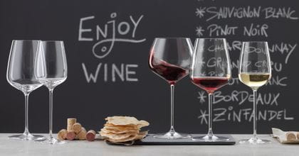 SPIEGELAU Willsberger Anniversary Gläser auf einer Marmoranrichte. Links ein leeres Bordeauxglas und ein Weißweinglas, in der Mitte ein Stapel Korken und Kartoffelchips, rechts das gefüllte Burgunderglas, das gefüllte Rotweinglas und das gefüllte Weißweinglas. Im Hintergrund eine Tafel mit einem Menü und der Aufschrift "Wein trinken".<br/>