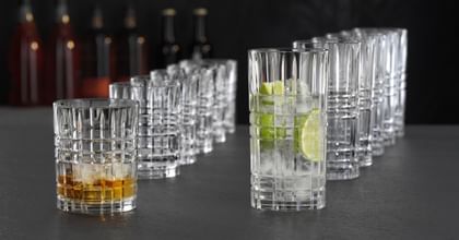 NACHTMANN Highland Square tumblers and longdrink glasses lined up in a row behind each other. The tumbler in front is filled with Whiskey on the rocks and the longdrink glass in front is filled with a clear drink with lime slices and ice cubes.