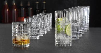 NACHTMANN Highland Square tumblers and longdrink glasses lined up in a row behind each other. The tumbler in front is filled with Whiskey on the rocks and the longdrink glass in front is filled with a clear drink with lime slices and ice cubes.<br/>