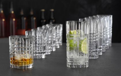 NACHTMANN Highland Square tumblers and longdrink glasses lined up in a row behind each other. The tumbler in front is filled with Whiskey on the rocks and the longdrink glass in front is filled with a clear drink with lime slices and ice cubes.<br/>