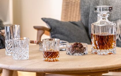 The NACHTMANN Noblesse tumbler with golden rim filled with whisky next to the bowl filled with snacks and the whisky decanter filled with whisky. All of them on a wooden table in front of a bench.<br/>
