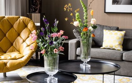 Die beeindruckenden, mit Blumensträußen gefüllten, NACHTMANN Minverva Vasen stehen auf runden Beistelltischen in einem Wohnzimmer. Im Raum befinden sich eine Couch, eine Lampe, ein Ledersessel und ein Teppich mit gelbem Muster.<br/>