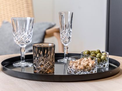 The NACHTMANN Noblesse collection on a serving tray. The smoke colored tumbler next to two Noblesse bowls, one filled with cashew nuts, one with olives. In the background are the Nobless wine glass and the Champagne glass.<br/>