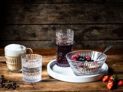 The NACHTMANN Ethno series on a wooden table. The hot beverage mug is filled with cappuccino, the tumbler with mineral water. The longdrink glass, filled with berry juice and the bowl, filled with a crunchy berry muesly, are standing on a white serving tray.<br/>