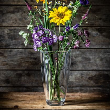 The crystal vase NACHTMANN Carre, filled with purple, white and yellow flowers on a wooden sideboard.<br/>