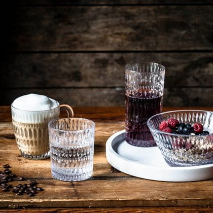 The NACHTMANN Ethno series on a wooden table. The hot beverage mug is filled with cappuccino, the tumbler with mineral water. The longdrink glass, filled with berry juice and the bowl, filled with a crunchy muesly, are standing on a white serving tray.<br/>