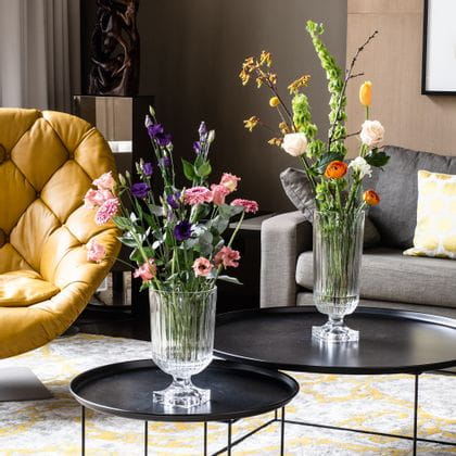 The impressive, with bouquets of flowers filled, NACHTMANN Minverva vases on round side tables in a living room. In the room there are a couch, a lamp, a leather armchair and a carpet with yellow pattern.<br/>
