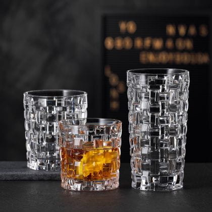 Il NACHTMANN Bossa Nova SOF riempito di whisky accanto al bicchiere longdrink Bossa Nova vuoto su una credenza in pietra nera. Dietro di loro il tumbler Bossa Nova vuoto.<br/>