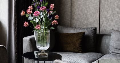 Die NACHTMANN-Vase Minerva mit platinbeschichtetem Fuß und einem großen Blumenstrauß auf einem runden Seitentisch. Im Hintergrund steht eine graue Couch mit einem braunen Kissen.<br/>