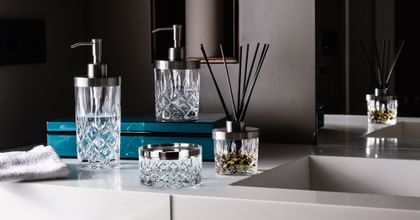 The NACHTMANN Spa Noblesse series on a bathroom sink in front of a mirror. The scent diffuser is filled with fragrance and black sticks. The small and the tall soap dispenser are filled with clear liquid. The storage jar is empty.