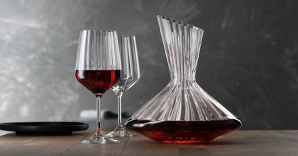 La carafe SPIEGELAU Lifestyle remplie de vin rouge sur une table. À gauche de la carafe, deux verres à vin rouge Lifestyle, dont l'un est rempli de vin rouge. Sur le bord gauche de la table, une assiette noire vide.<br/>