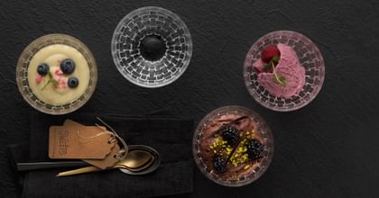 Four NACHTMANN Bossa Nova dip bowls, one filled with custard and blueberries on top, one with ice cream and a raspberry on top, one with chocolate cream with blackberries on top and one empty. To the right of it are spoons. All of them are on a black plate