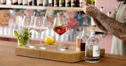 Vasos de Gin Tonic NACHTMANN con pajitas de cristal sobre la barra de un bar. Uno está lleno de un Gin Cocktail rojo con menta y cáscara de limón. Detrás de los vasos hay un limón y un vaso con ramitas de menta. A la derecha, las manos del camarero y el equipo de bar.<br/>