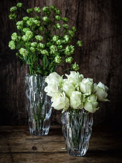 The small oval NACHTMANN Quartz vase filled with white roses in front of the tall NACHTMANN Quartz vase filled with white flowers on a sideboard made of dark wood.<br/>