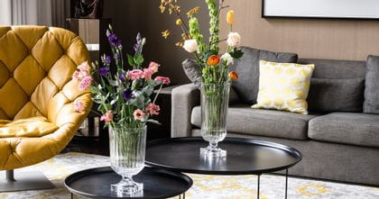 The impressive, with bouquets of flowers filled, NACHTMANN Minverva vases on round side tables in a living room. In the room there are a couch, a lamp, a leather armchair and a carpet with yellow pattern.<br/>
