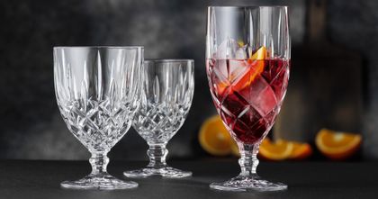 Das eisgekühlte NACHTMANN Noblesse-Glas, gefüllt mit einem roten Cocktail, Eiswürfeln und einer Orangenscheibe, neben dem leeren großen und dem leeren kleinen Kelch. Im Hintergrund sind Orangen zu sehen.<br/>