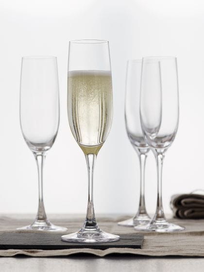 Four SPIEGELAU Vino Grande Champagne flutes on a table with a tablecloth. One glass, filled with Champagne, is standing in the foreground on a slate coaster.<br/>