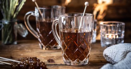 The NACHTMANN Noblesse tea mug with hot tea in it and rock candy. On the wooden table, there are more sticks with brown rock candy, a jar with lavender flowers, a second filled crystal tea mug and a shining tea light.<br/>