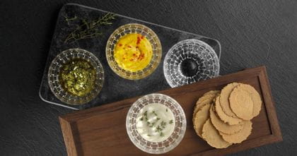 Four NACHTMANN Bossa Nova dip bowls, three of them are filled with different dips and one is empty. Three bowls are on a small glass plate. One filled bowl is on a wooden board with cookies next to it.<br/>
