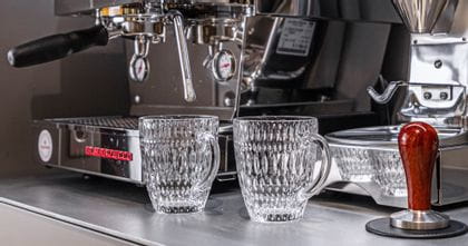 Deux tasses à boissons chaudes NACHTMANN Ethno vides devant une machine à portafilter et un moulin à café. Sur la machine, d'autres tasses à café Ethno sont en train de chauffer. À droite, un tamper à café.<br/>