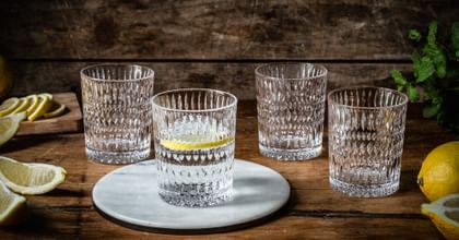Four NACHTMANN Ethno tumblers on a wooden sideboard. One of them is filled with water and a slice of lemon and stands on a round stone platter. Around the tumblers are lemons, slices of lemon, and mint sprigs.<br/>