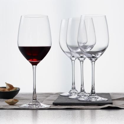 Vier SPIEGELAU Vino Grande Rotweingläser auf einem Tisch mit einem Tischtuch. Das linke Glas ist mit Rotwein gefüllt, links davon eine kleine Schale mit Keksen.<br/>