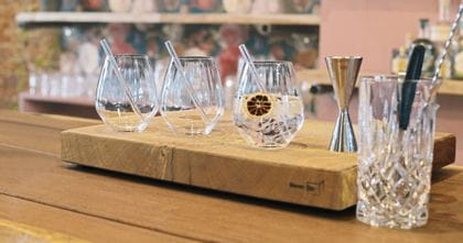 Drei NACHTMANN Gin und Tonic Gläser aus der Serie Tastes Good. Jedes hat einen Glasstrohhalm im Inneren, eines ist mit einem Gin-Cocktail gefüllt. Rechts neben den Gläsern steht ein NACHTMANN Noblesse Rührglas und Barausstattung.<br/>