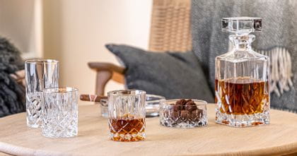 El vaso NACHTMANN Noblesse con borde dorado lleno de whisky junto al cuenco lleno de aperitivos y la jarra llena de whisky. Todos ellos sobre una mesa de madera frente a un banco.<br/>