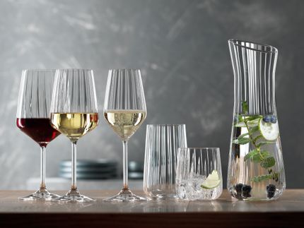 Die SPIEGELAU Lifestyle-Serie auf einem Tisch. Das gefüllte Rotweinglas, das gefüllte Weißweinglas, das gefüllte Champagnerglas, ein leeres Longdrinkglas, ein Becher gefüllt mit Wasser, Eiswürfeln und einer Limettenscheibe. Rechts die Karaffe mit gefülltem Wasser.<br/>