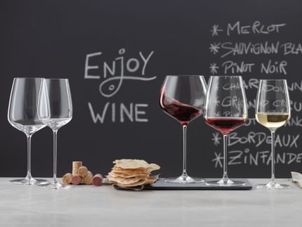 SPIEGELAU Willsberger Anniversary Gläser auf einer Marmoranrichte. Links ein leeres Bordeauxglas und ein Weißweinglas, in der Mitte ein Stapel Korken und Kartoffelchips, rechts das gefüllte Burgunderglas, das gefüllte Rotweinglas und das gefüllte Weißweinglas. Im Hintergrund eine Tafel mit einem Menü und der Aufschrift "Wein trinken".<br/>