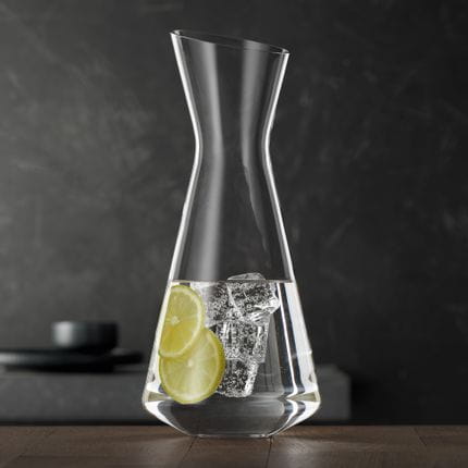 La esbelta jarra SPIEGELAU Style llena de agua, rodajas de limón y cubitos de hielo sobre una mesa de madera.<br/>