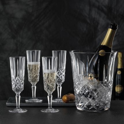 Vier NACHTMANN Noblesse Champagnergläser, von denen zwei mit Champagner gefüllt sind. Daneben steht der NACHTMANN Noblesse Champagner-Kühler mit Eiswürfeln und einer offenen Flasche Champagner.<br/>