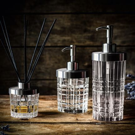The NACHTMANN Spa Square series on a wooden sideboard. The scent diffuser is filled with a fragrance and eight black sticks. To the right are the small and the tall soap dispenser.<br/>