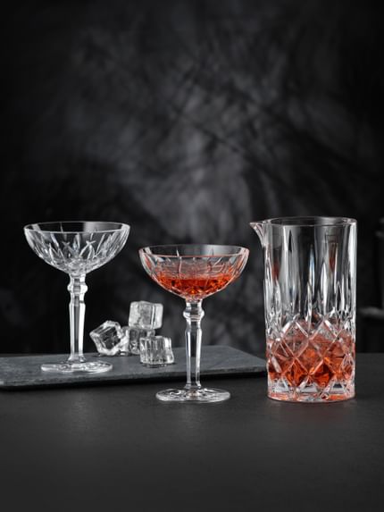 Der NACHTMANN Noblesse-Krug, gefüllt mit einem roten Getränk und Eiswürfeln, und zwei NACHTMANN Noblesse-Cocktailgläser, eines davon gefüllt mit demselben roten Getränk. Im Hintergrund ist das leere Cocktailglas mit Eiswürfeln zu sehen.<br/>