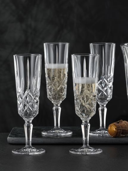 Four NACHTMANN Noblesse Champagne glasses two of which are filled with Champagne. Next to them is the NACHTMANN Noblesse Champagne cooler with ice cubes and an open bottle of Champagne inside.<br/>