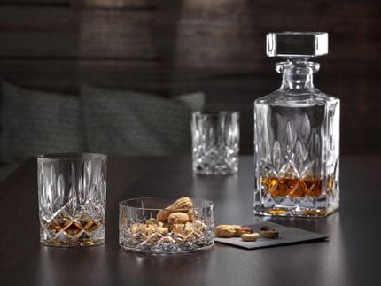 The NACHTMANN Noblesse tumbler filled with whisky next to the snack bowl filled with peanuts. In the background is the NACHTMANN Noblesse crystal whisky decanter filled with whisky.<br/>