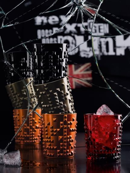 Vier NACHTMANN Punkbecher in Tiefschwarz, Kupfer und Gunmetal vor einem zerbrochenen Spiegel aufgestapelt. Daneben der Punkbecher in Rubinrot, gefüllt mit Eiswürfeln. Auf dem Spiegel ist ein Union Jack und die Zeile nextgen goes punk zu sehen.<br/>