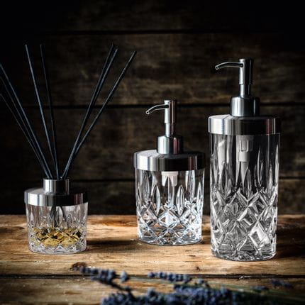 The NACHTMANN Spa Noblesse series on a wooden sideboard. The scent diffuser is filled with a fragrance and eight black sticks. To the right are the small and the tall soap dispenser.<br/>
