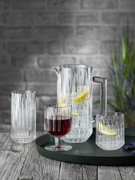 The NACHTMANN Jules crystal glass series on a round serving tray. The pitcher and the tumbler are filled with water, ice cubes and lemon slices, to their right is lavender. The goblet is filled with red wine. The longdrink glass in the background is empty.<br/>