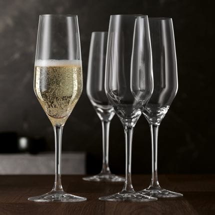 Four SPIEGELAU Style Champagne flutes on a wooden table. One glass is filled with sparkling wine.<br/>