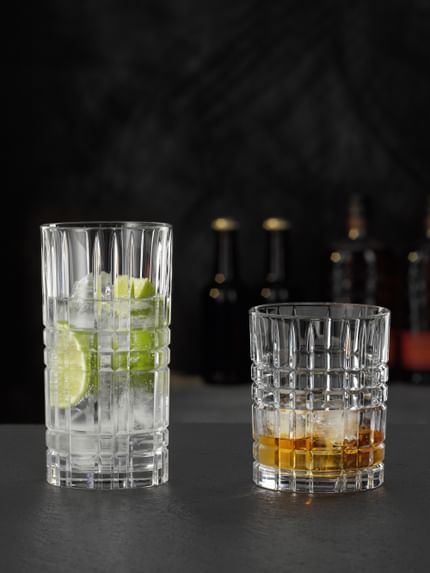 Das NACHTMANN Highland Square Longdrinkglas gefüllt mit einem klaren Drink mit Limette und Eiswürfeln und der Tumbler gefüllt mit Whiskey auf Eis.<br/>