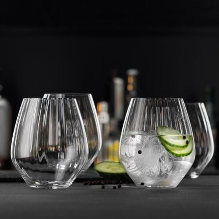 Four SPIEGELAU Gin and Tonic tumblers with optical line effects in the glass design on a table. One of the glasses is filled with a Gin and tonic cocktail with cucumber and black pepper.<br/>