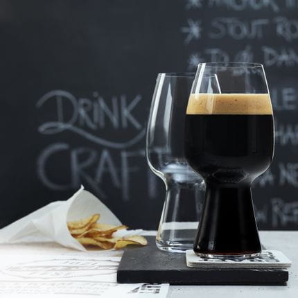 Zwei SPIEGELAU Craft Beer Gläser für Stout auf einem Schiefertablett. Eines davon ist mit dunklem Stout-Bier gefüllt, das Glas dahinter ist leer. Dahinter steht eine kleine Tüte mit Kartoffelchips und eine Tafel mit einer Bierkarte darauf.<br/>