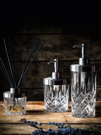 The NACHTMANN Spa Noblesse series on a wooden sideboard. The scent diffuser is filled with a fragrance and eight black sticks. To the right are the small and the tall soap dispenser.<br/>