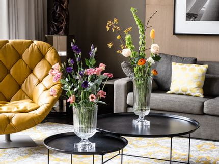 L'impressionnant vase Minverva de NACHTMANN, rempli de bouquets de fleurs, est posé sur des tables d'appoint rondes dans un salon. La pièce comprend un canapé, une lampe, un fauteuil en cuir et un tapis à motifs jaunes.<br/>
